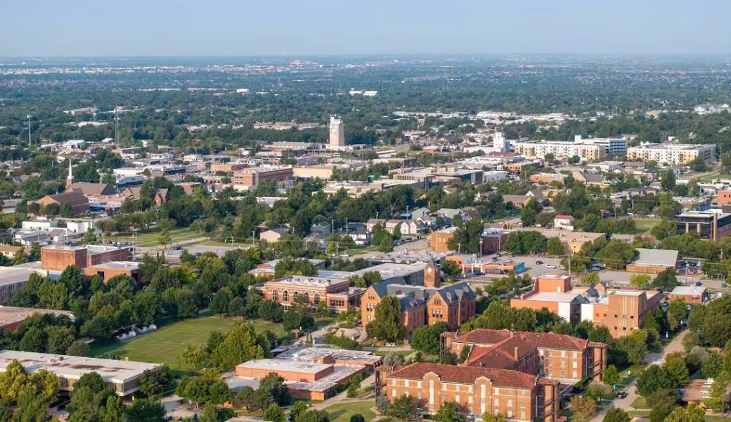 Aerial view of Norman, OK, featuring a mix of residential neighborhoods, tree-lined streets, and campus-style buildings, with a vibrant cityscape and surrounding greenery stretching into the distance.