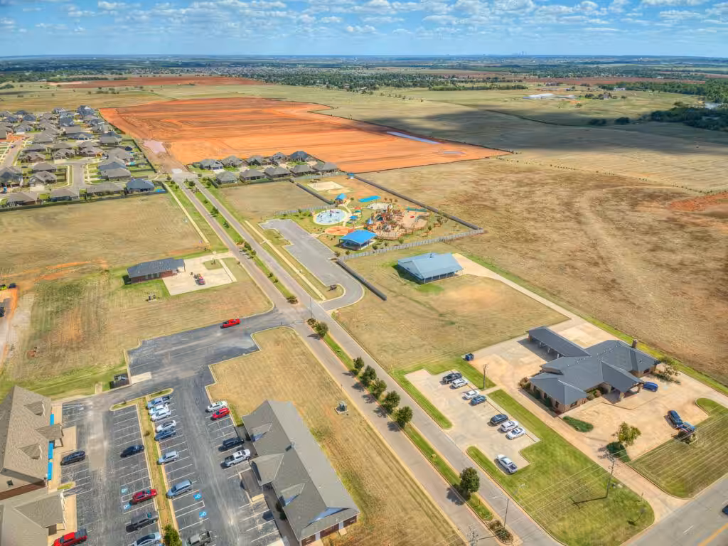 Aerial view of a residential area in Piedmont, OK, featuring a neighborhood park with playground equipment, nearby homes, open fields, and quiet suburban streets.
