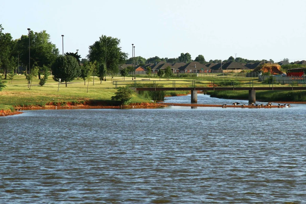 Park scene in Mustang, OK, featuring a calm pond with ducks, a small pedestrian bridge, green open space, and nearby residential homes in the background.